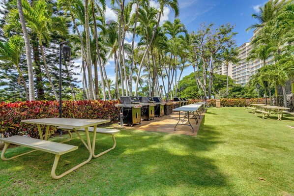 A grassy outdoor area with picnic tables and barbecue grills, surrounded by palm trees and a building in the background. - 1 Mi to Makaha Beach! Resort Escape w/ Pool Access (Waianae)