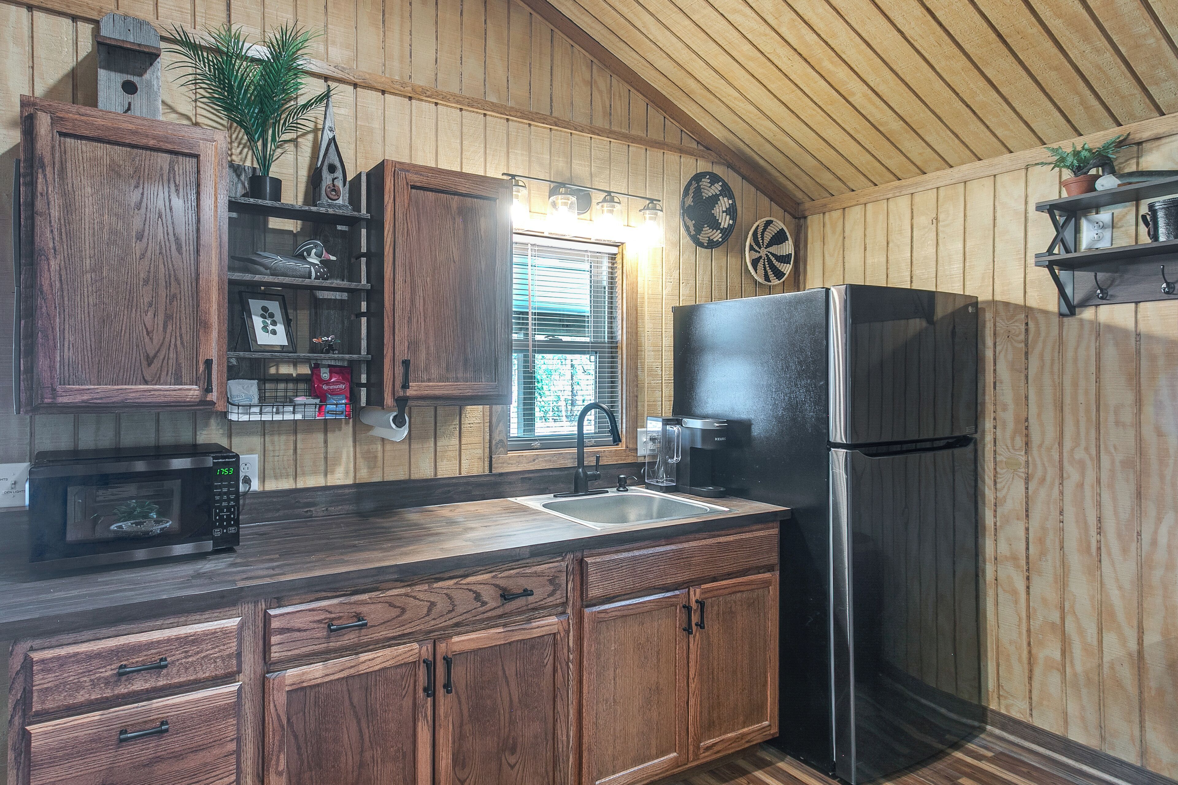 Une cuisine avec des meubles de rangement en bois, un four à micro-ondes, un réfrigérateur, un évier et une fenêtre avec des stores.