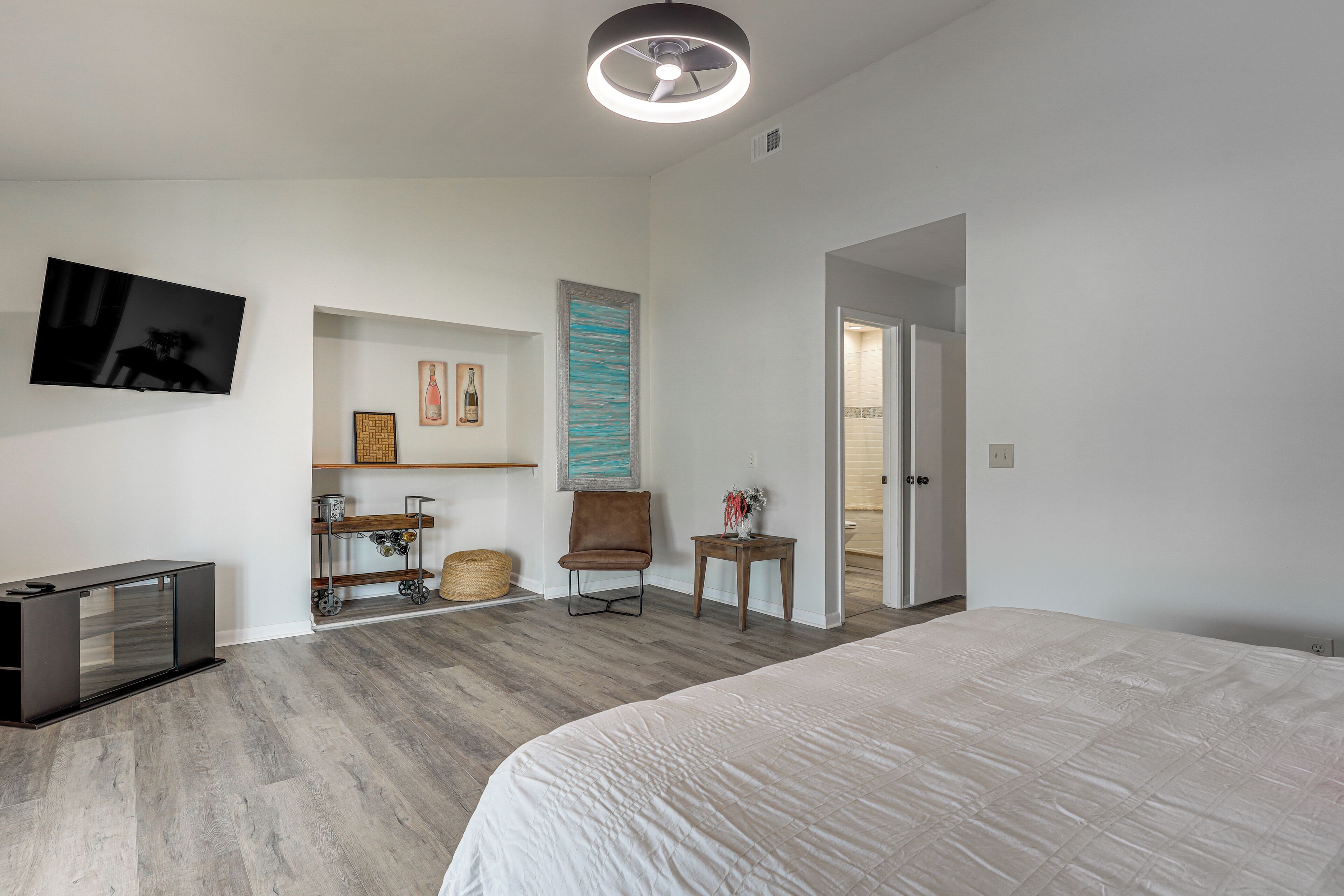 A bedroom with a bed, a TV, a chair, a small table, and a door leading to another room.