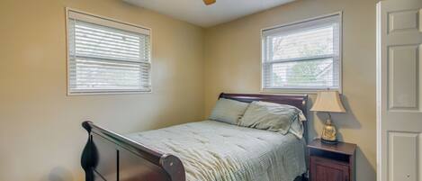 A bedroom with a bed, nightstand, lamp, ceiling fan, and two windows.