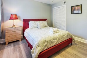 Ein ordentlich gemachtes Bett mit rotem Kopfteil, eine Nachttischlampe, eine Holzkommode und ein weißer Kleiderschrank mit einem Bild an der Wand.