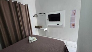 A hotel room with a bed, a TV mounted on the wall, a shelf with a remote control, and a small wall-mounted rack.