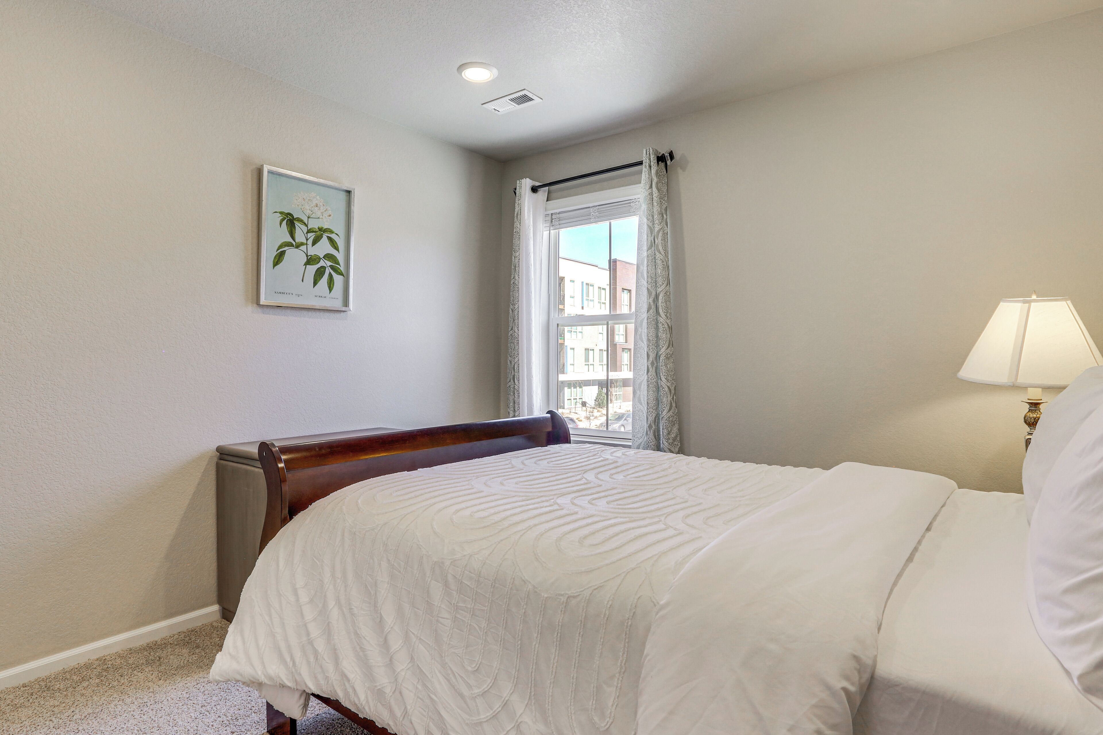 A bedroom with a bed, a window with curtains, a wall-mounted picture, and a bedside lamp.