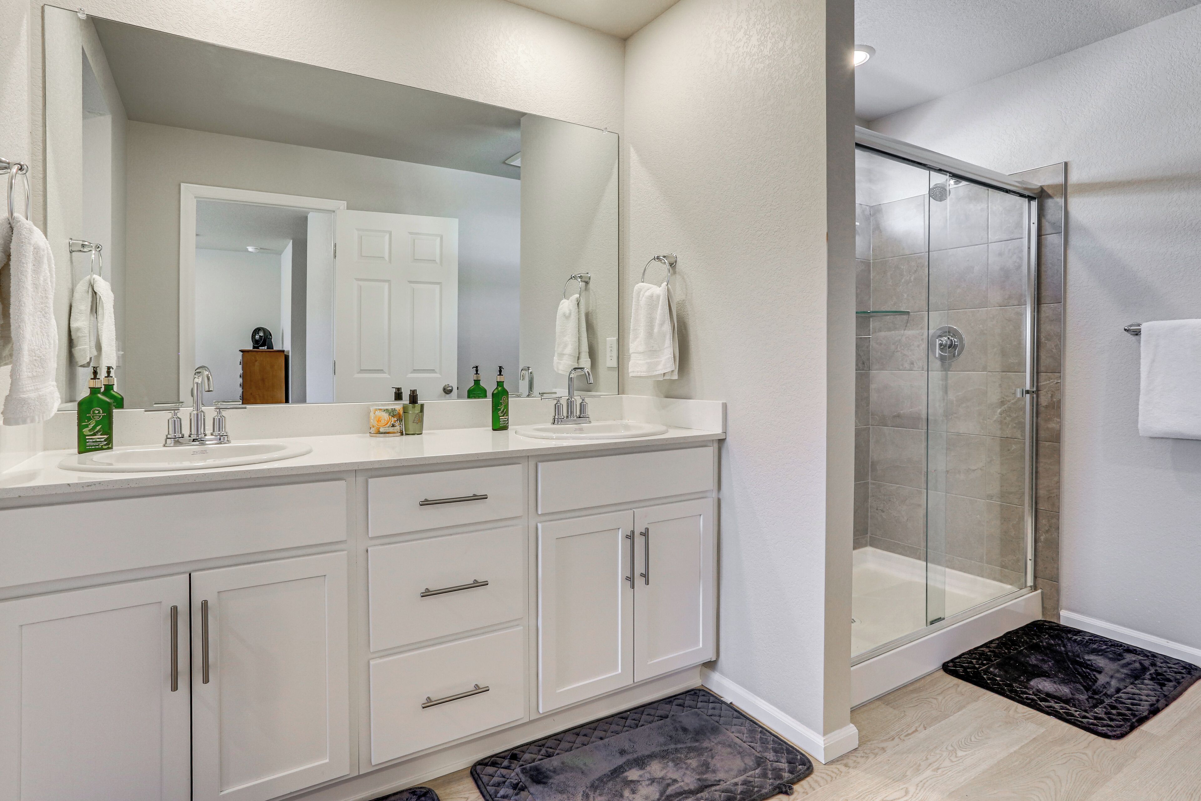 A bathroom with a large mirror, two sinks, a shower, and a black bath mat.