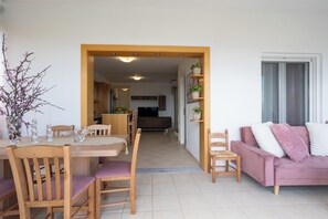 A dining area with wooden chairs and a table, a pink sofa, and a kitchen in the background. - Medusa Panoramic View Apartment in Georgioupolis (96FW+GW Exopoli)