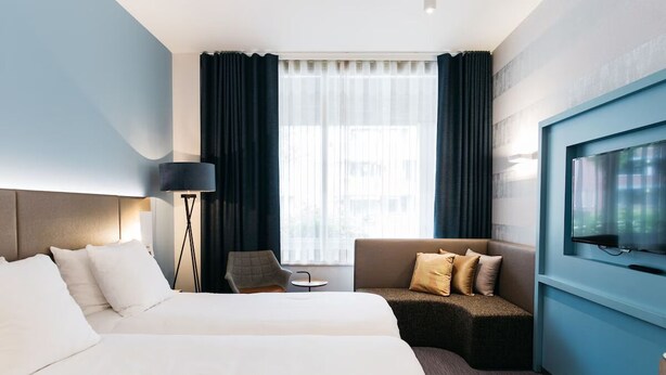 Une chambre d’hôtel avec deux lits, un canapé, un fauteuil, une télévision et une fenêtre avec des rideaux.