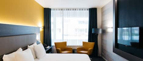Ein Hotelzimmer mit einem großen Bett, zwei Sesseln, einem Fernseher und einem Fenster mit Vorhängen.