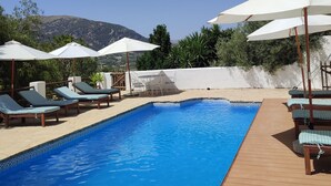 Una piscina con sillones de descanso y sombrillas, rodeada por una terraza de madera y un muro blanco.