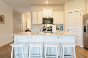 A kitchen with white cabinets, a stainless steel refrigerator, a microwave, and a sink. There are four white stools at the island. - Family-Focused Beach Bungalow Steps from the Beach with Resort Pool (St Teresa)