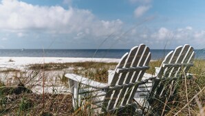 Two Adirondack chairs facing a beach with sand and grass, under a partly cloudy sky.