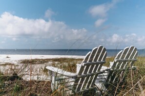 Two Adirondack chairs facing a beach with sand and grass, under a partly cloudy sky. - Family-Focused Beach Bungalow Steps from the Beach with Resort Pool (St Teresa)