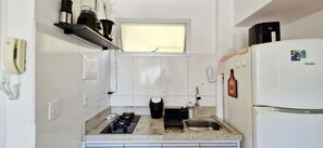 Une cuisine de taille réduite, équipée d’une cuisinière à gaz, d’un évier, d’un réfrigérateur et d’une fenêtre avec des stores.