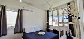 Habitación de hotel con cama, ventana con cortinas, unidad de aire acondicionado, ventilador de techo y vista de la ciudad.