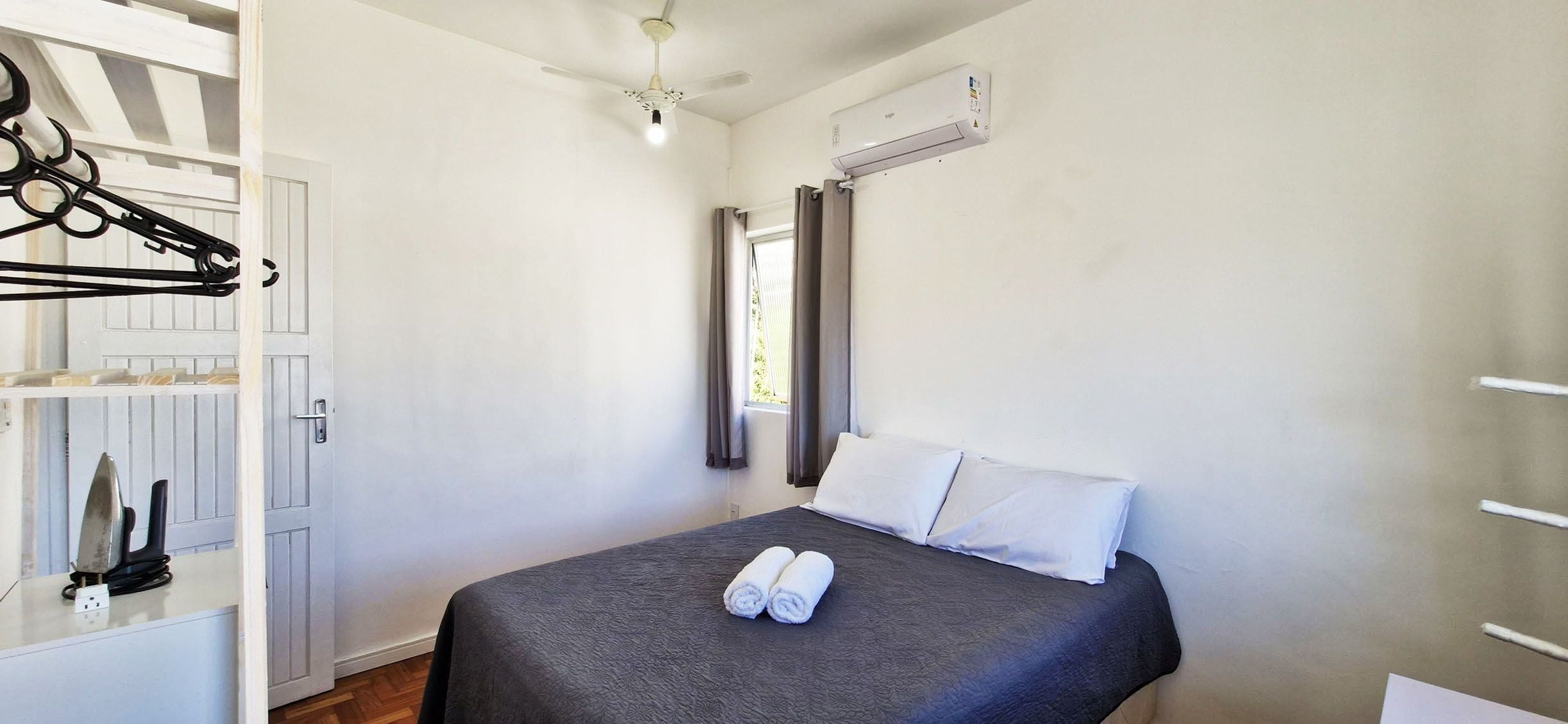 A hotel room with a bed, a ceiling fan, an air conditioning unit, a window with curtains, and a towel on the bed.