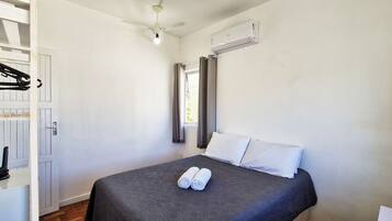 A hotel room with a bed, a ceiling fan, an air conditioning unit, a window with curtains, and a towel on the bed.