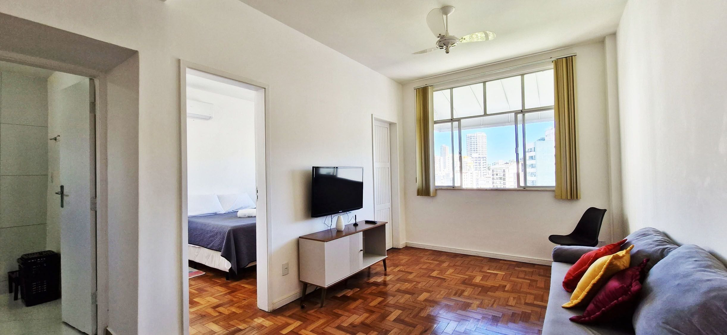 A room with a bed, a TV, a window with curtains, a ceiling fan, and a herringbone-patterned floor.