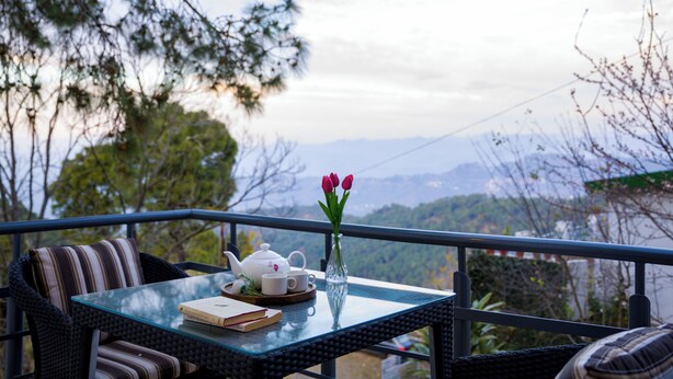 A balcony with a table, a wicker chair, a glass vase with flowers, and a view of a mountainous landscape. - Alaya Stays Mehar Villa 3BHK With Garden (Kasauli)