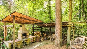 A rustic outdoor kitchen with a chimney, wooden benches, and a covered seating area surrounded by trees. - Brechts Boshuisje Quiet Holiday Home with Sauna (Brecht)