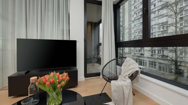 Ett modernt vardagsrum med en stor platt-TV, en svart soffa, en glasvas med tulpaner och utsikt över en stadssiluett genom stora fönster.