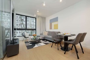 A modern living room with a sofa, dining table, and chairs. There is a TV on a stand, a coffee table with flowers, and a view of buildings through large windows. - Spacious Apartment in the Center | Workspace (Warszawa)