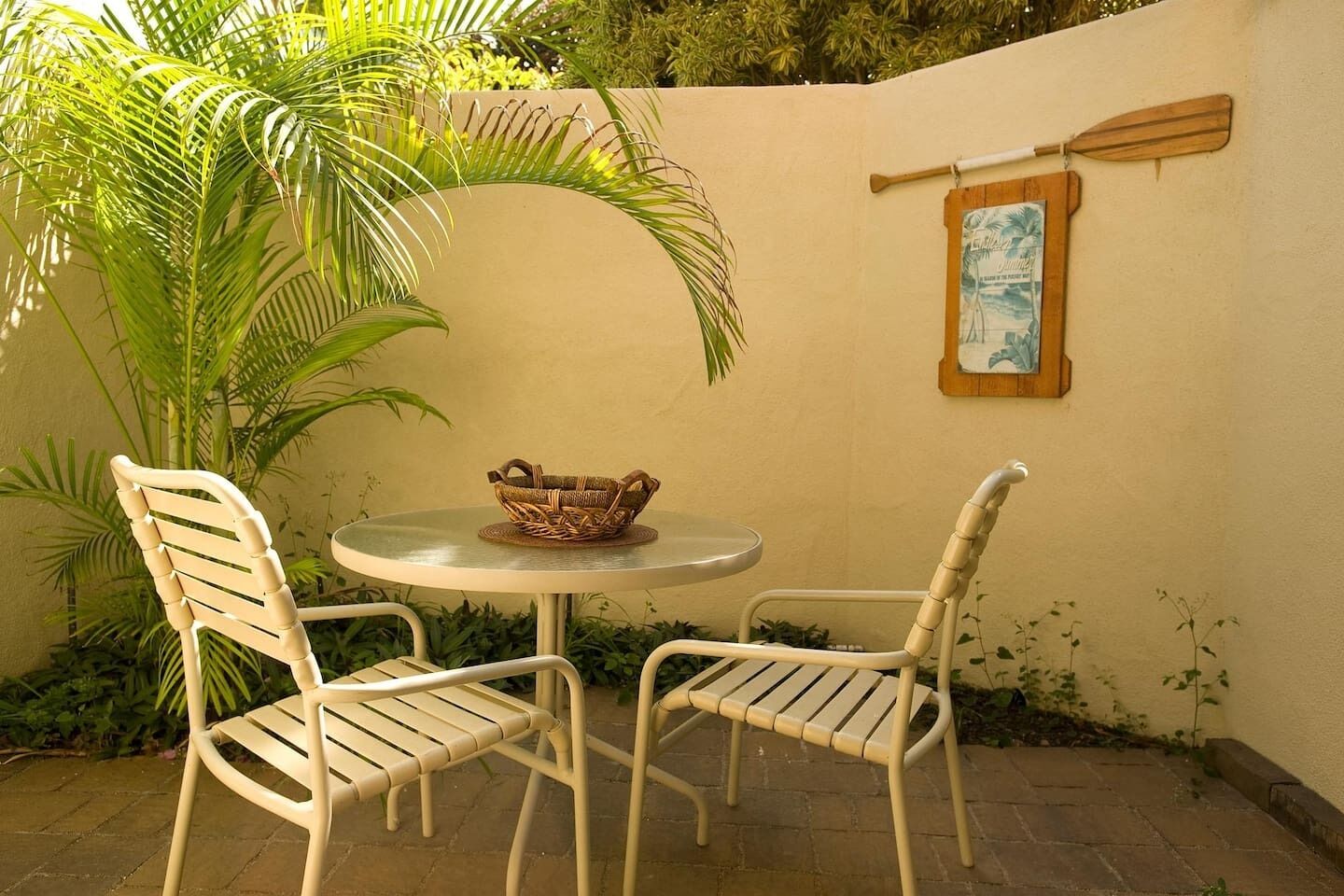 Eine Terrasse mit zwei weißen Metallstühlen, einem runden Tisch, einem gewebten Korb und einem Holzschläger an der Wand.