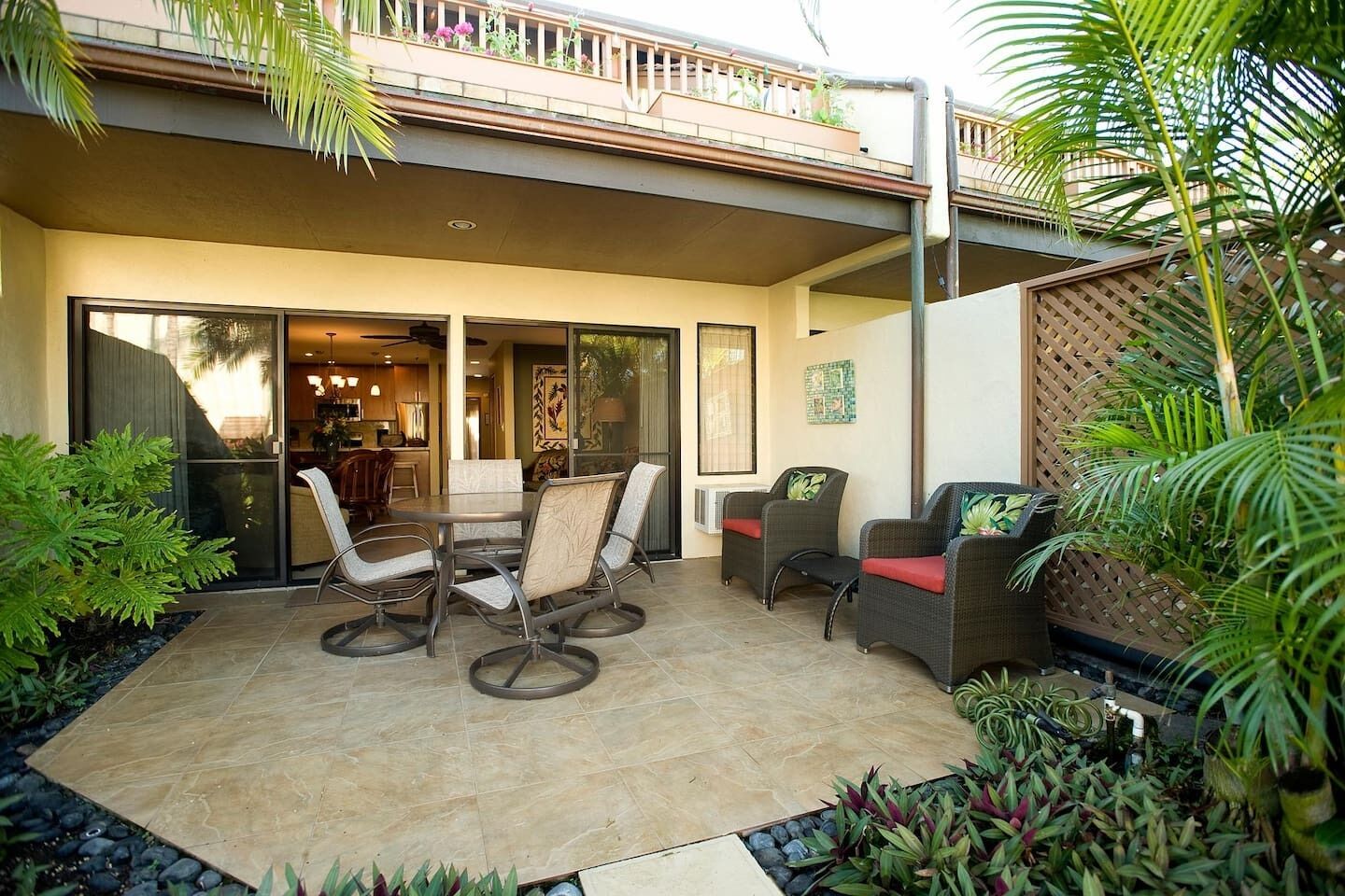 A patio with a table and chairs, a balcony with plants, and a sliding glass door leading to a kitchen.