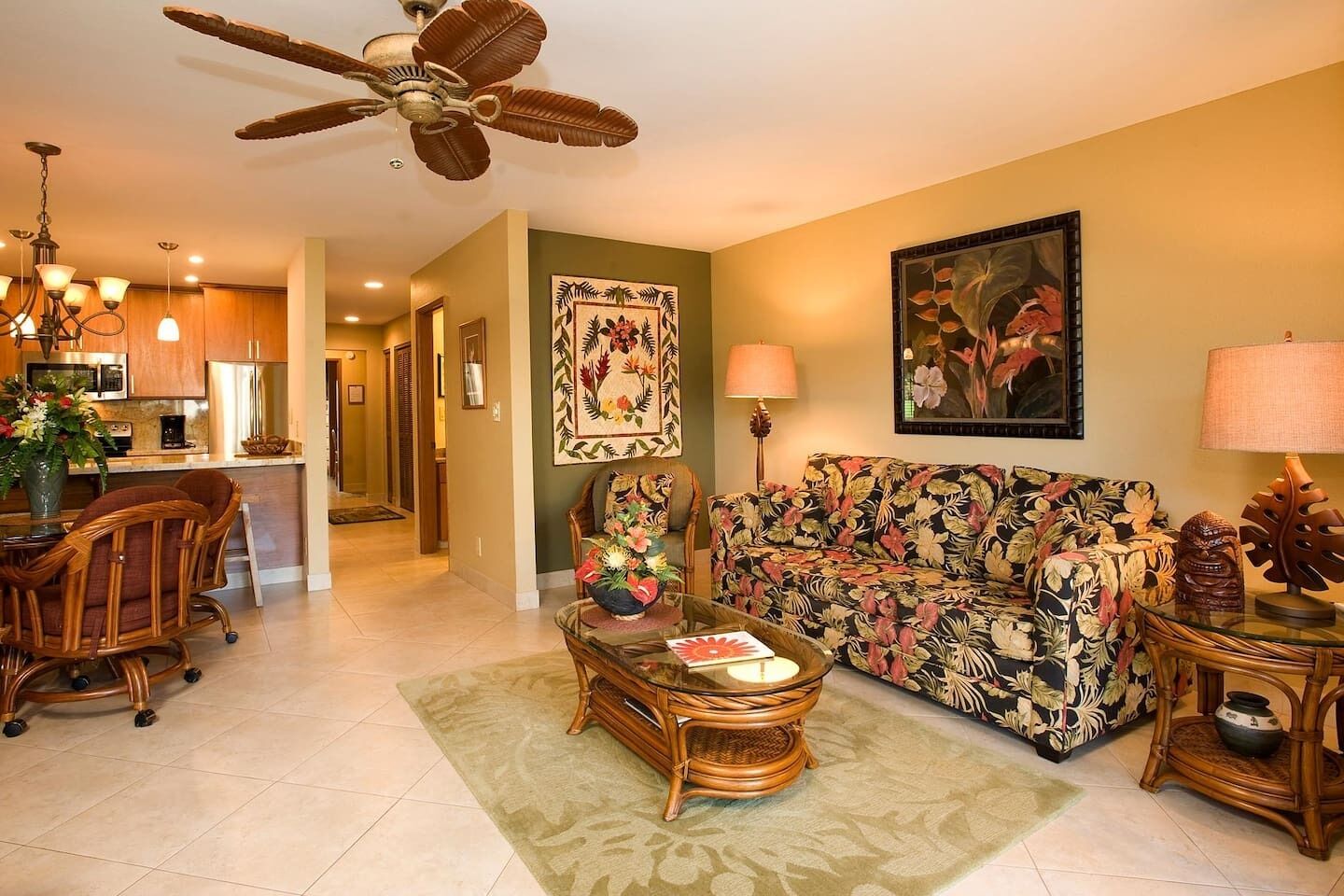A living room with a sofa, chairs, a coffee table, a ceiling fan, and a kitchen area in the background.