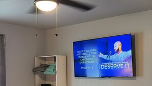 Un dormitorio con una cama, ventilador de techo, televisor en la pared y estantería.