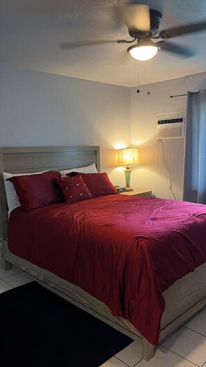 Une chambre à coucher comprenant un lit, une table de chevet, une lampe, un ventilateur de plafond et un appareil de climatisation.