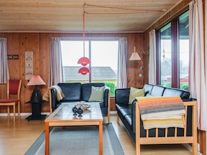 A cozy living room with a wooden ceiling, a sofa, a coffee table, and a view through large windows.
