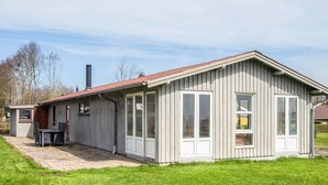 Ein eingeschossiges Gebäude mit geneigtem Dach, großen Fenstern und einer kleinen Terrasse mit Tisch und Stühlen.
