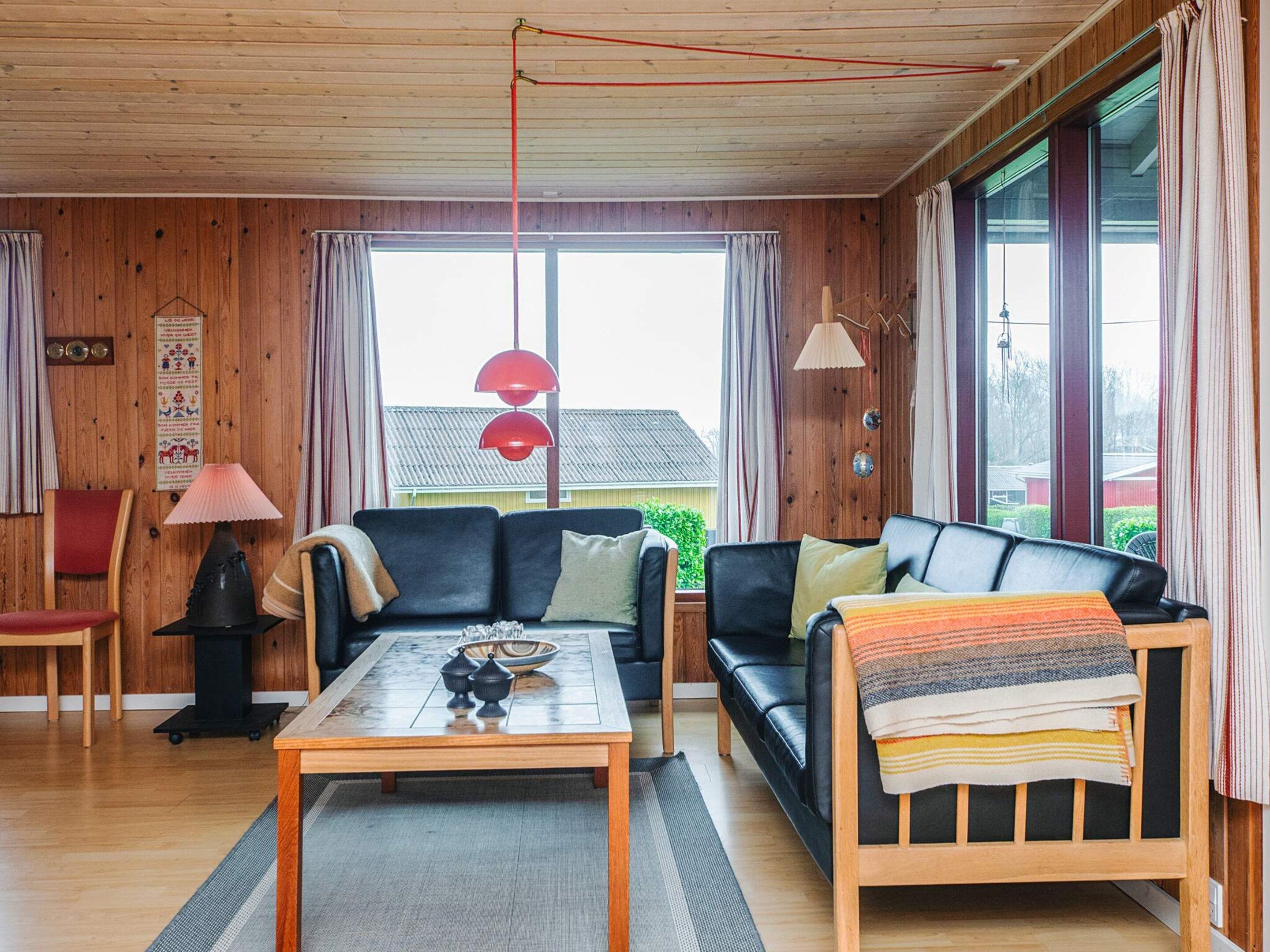 Een gezellige woonkamer met een houten plafond, een zwarte leren bank, een houten salontafel en een rode hanglamp.