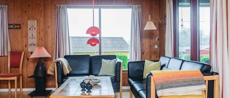 Een gezellige woonkamer met een houten plafond, een zwarte leren bank, een houten salontafel en een rode hanglamp.