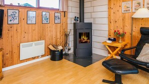Ein gemütlicher Raum mit Holzwandverkleidung, einem schwarzen Holzofen, einer Heizung, einem Sessel und gerahmten Pflanzenbildern an der Wand.