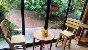 Ein Zimmer mit Holztisch und zwei Stühlen, einer Vase mit gelben Blumen sowie Blick auf einen Ziegelweg und Grünflächen draußen.