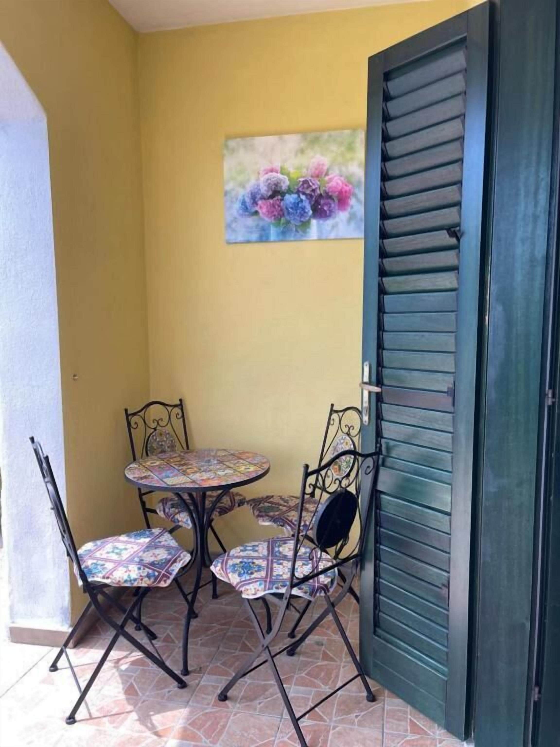 A small patio with a round table and four chairs, a door with horizontal slats, and a floral painting on the wall.