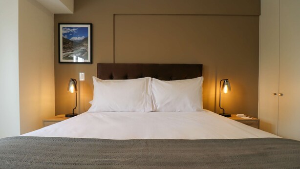 Een net opgemaakt bed met een hoofdbord, twee bedlampjes en een lijst aan de muur.