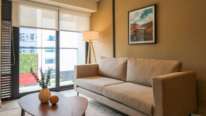 Een moderne hotelkamer met een beige bank, een tafel met twee vazen, een vloerlamp en een groot raam met jaloezieën.