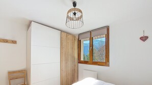 Quarto com cama, cadeira de madeira, guarda-roupa, janela com vista e luminária pendente.