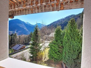 A mountain view from a room with a wooden ceiling, a window overlooking a road with houses, and a mountain range in the distance. - Snow 19 – Sleeps 5 – Cosy, bright apartment with mountain views – Saint Jean D'Aulps – Near Morzine (Saint-Jean-d´Aulps)