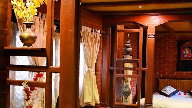A room with a bed, a table with a lace tablecloth, a wooden shelf with a decorative item, and a window with curtains. - 
Charming Newari-modern homestay in Patan, cozy stay near heritage sites (Lalitpur)