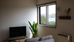 A room with a bed, a TV on a stand, a potted plant, a window with a view, and a radiator. - YORIDOKORO (Oma)