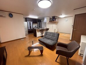 A modern living room with a sofa, armchair, and glass coffee table. There is a kitchen area with cabinets and appliances in the background.