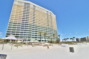 A high-rise hotel with multiple balconies, a beach area with palm trees, and a clear blue sky. - Gulf Views & Family Fun – 3BR Boardwalk Condo (Panama City Beach)