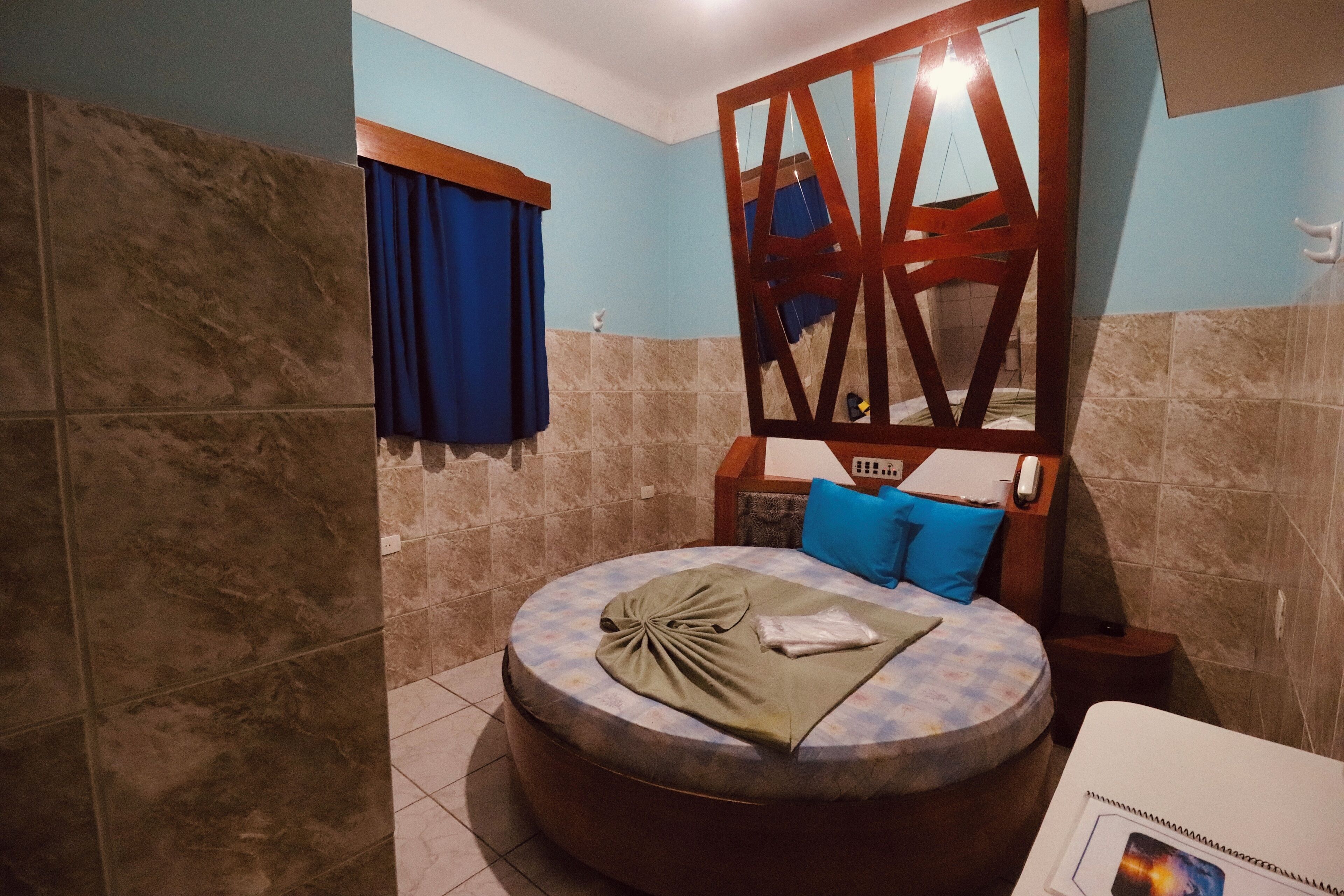 Een rond bed met een blauw-witte bedspreidek, een houten hoofdbord en een klein nachtkastje met een afstandsbediening.