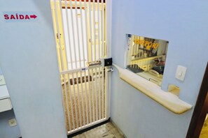 A hotel room with a white door marked 'SAÍDA' (exit), a reception area with a 'RECEPCÃO' sign, and a small window with a view of the interior. - Hotel Bellamy (São Paulo)