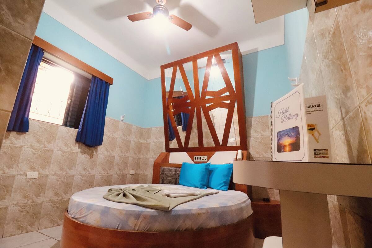 A hotel room with a bed, a wooden headboard, a window with curtains, a ceiling fan, and a wall-mounted TV.
