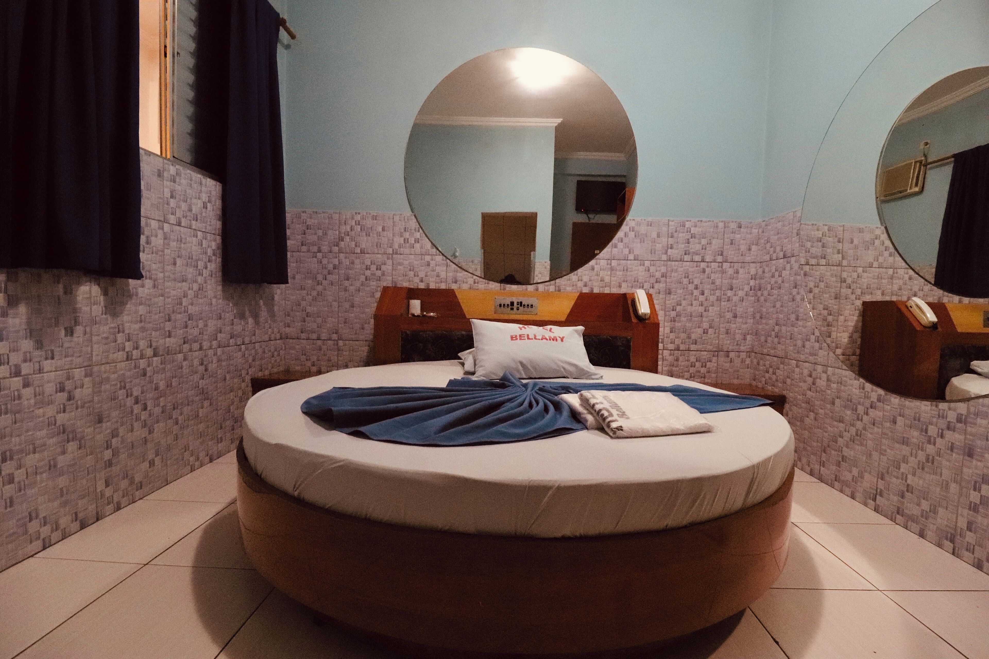 Ein rundes Bett mit weißer Matratze, einer blauen Decke und einem Kissen mit dem Aufdruck 'BELLAMY' in einem Raum mit gefliesten Wänden und einem Spiegel über dem Bett.