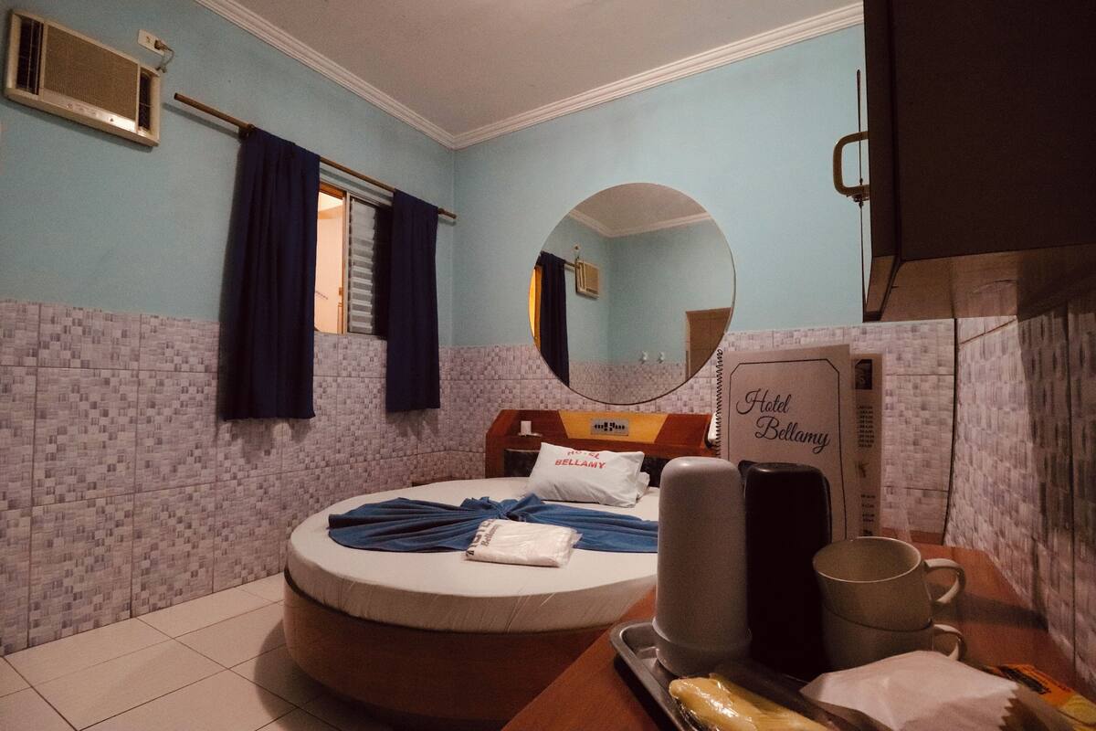 A hotel room with a bed, a round mirror, a bedside table with a lamp, a wall-mounted air conditioner, and a sign indicating the hotel name.