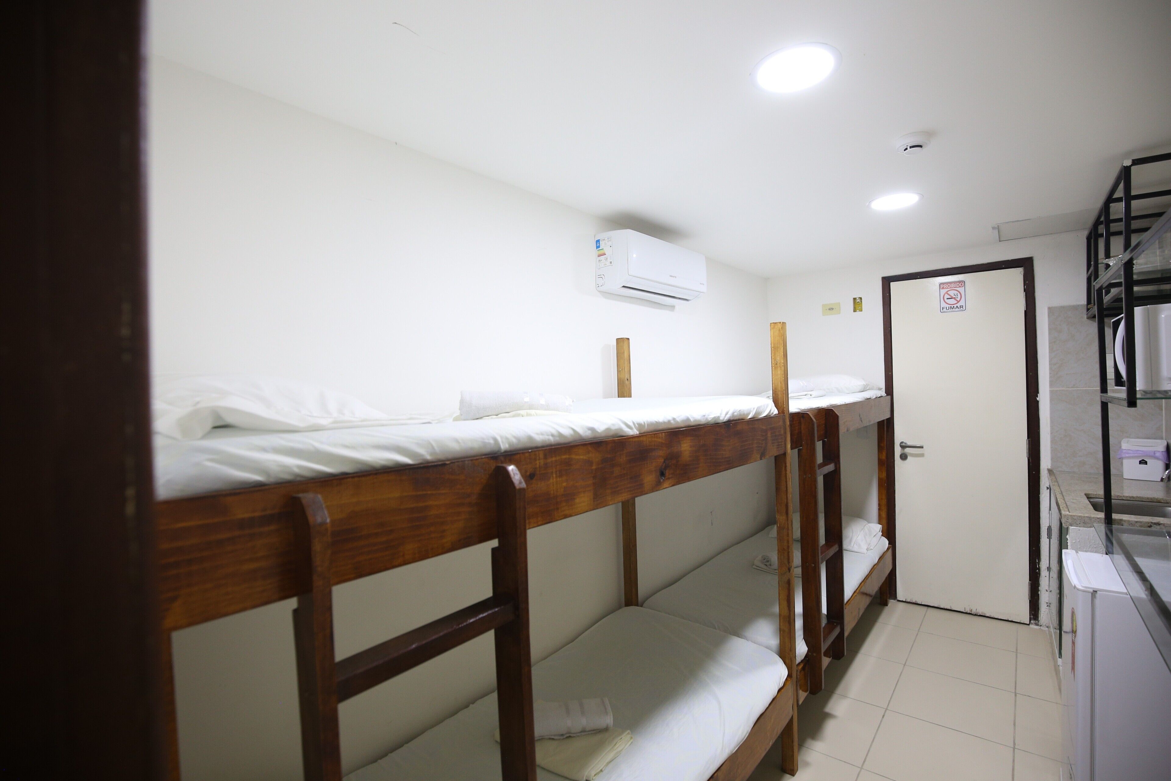 Une chambre de dortoir avec des lits superposés, un climatiseur blanc, une porte sur laquelle est apposée une pancarte « Il est interdit de fumer » et un petit coin cuisine.